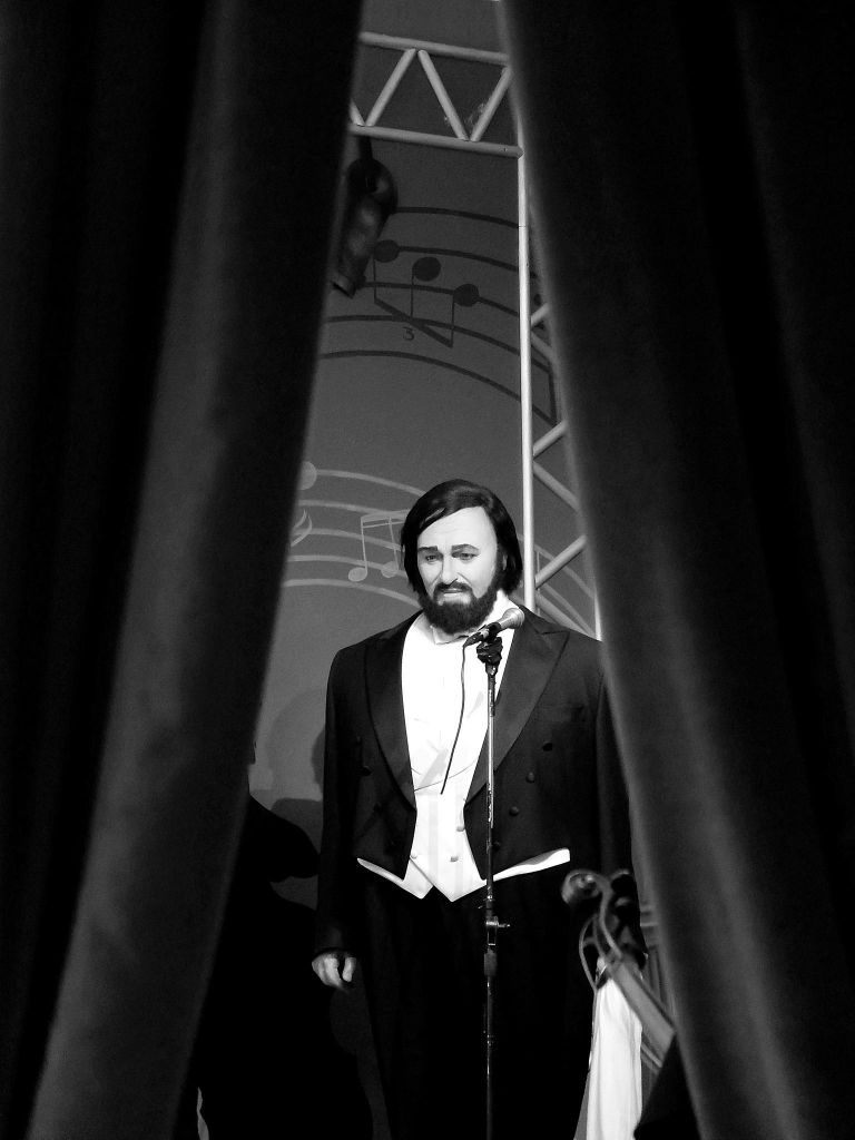 정장 차림의 남성이 마이크를 들고 배경에 악보가 있는 커튼 사이에 서 있습니다. 흑백 사진.