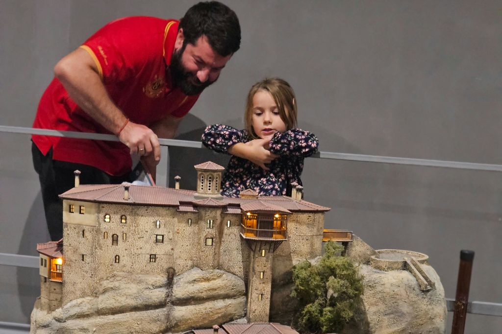 수염을 기른 남자와 어린 소녀가 바위가 많은 풍경에서 고대 건물의 상세한 미니어처 모델을 바라보고 있습니다.
