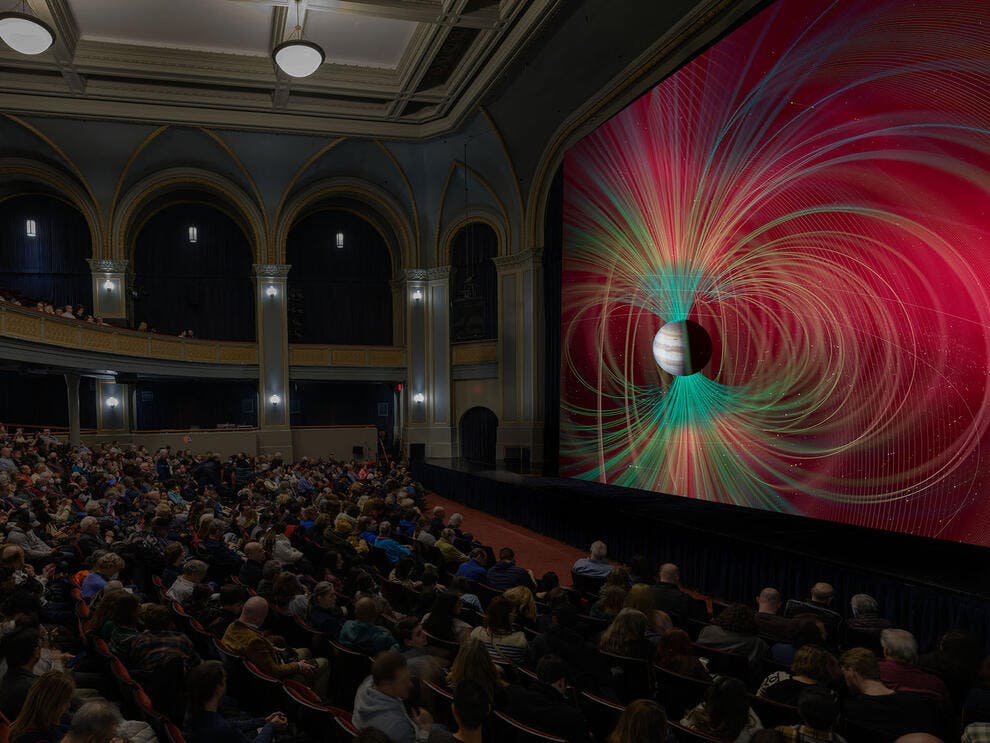 소용돌이 치는 선이있는 행성을 포함하여 다채로운 공간 이미지를 표시하는 대형 스크린을 시청하는 극장 관객이 붐빕니다.