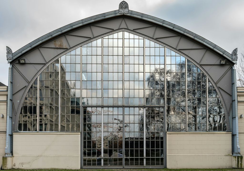 나무와 다른 구조물의 반사를 보여주는 반사 유리 패널로 완전히 덮인 커다란 아치형 건물.