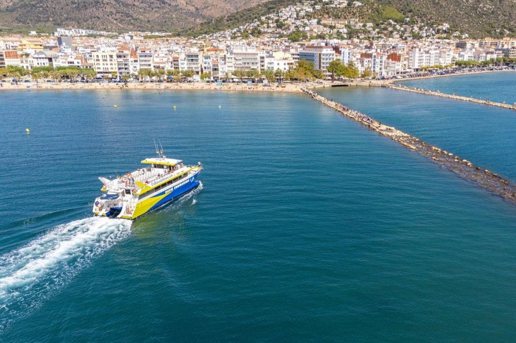 건물이 줄지어 있고 산악 지대를 배경으로 해안 마을 근처의 잔잔한 푸른 바다에서 항해하는 페리보트.