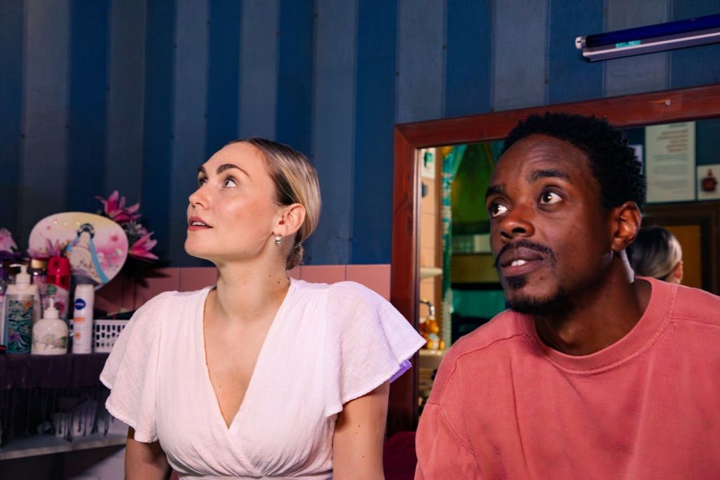 흰색 상의를 입은 여성과 분홍색 셔츠를 입은 남성이 배경에 파란색 벽이 있는 실내에서 세심하게 카메라를 벗어난 모습을 하고 있습니다.