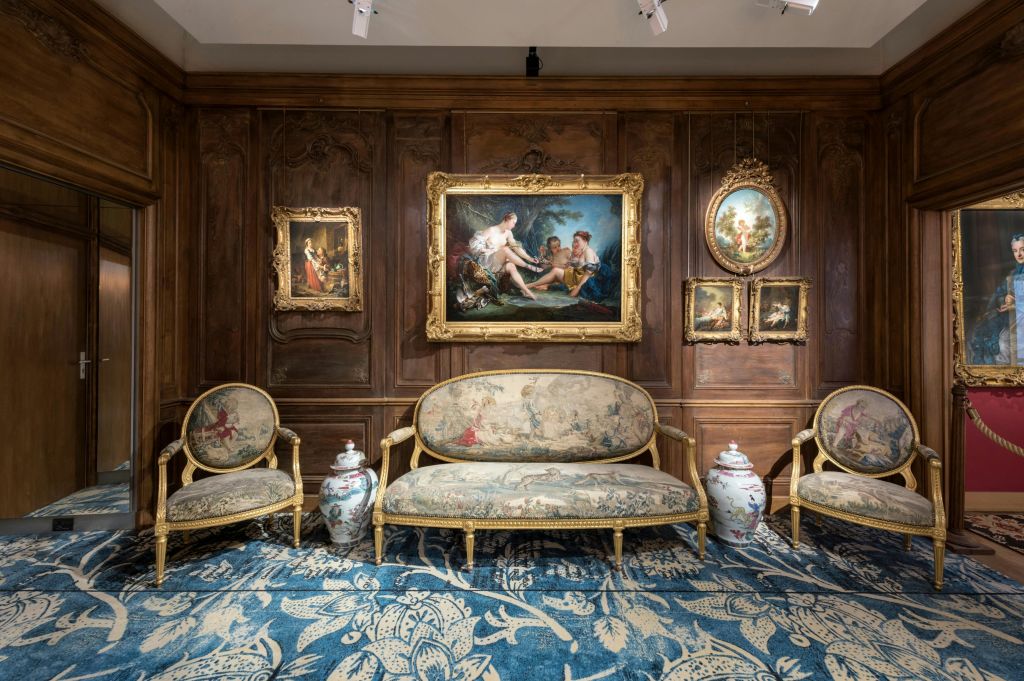 나무 패널 벽에 무늬가 있는 파란색 러그 위에 앤티크 가구, 액자 그림, 장식용 꽃병이 놓인 화려한 거실.