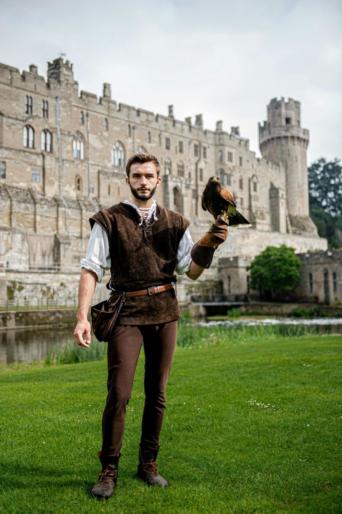 중세 복장을 한 한 남성이 탑과 주변 녹지가 있는 커다란 석조 성 앞에서 매를 들고 있습니다.