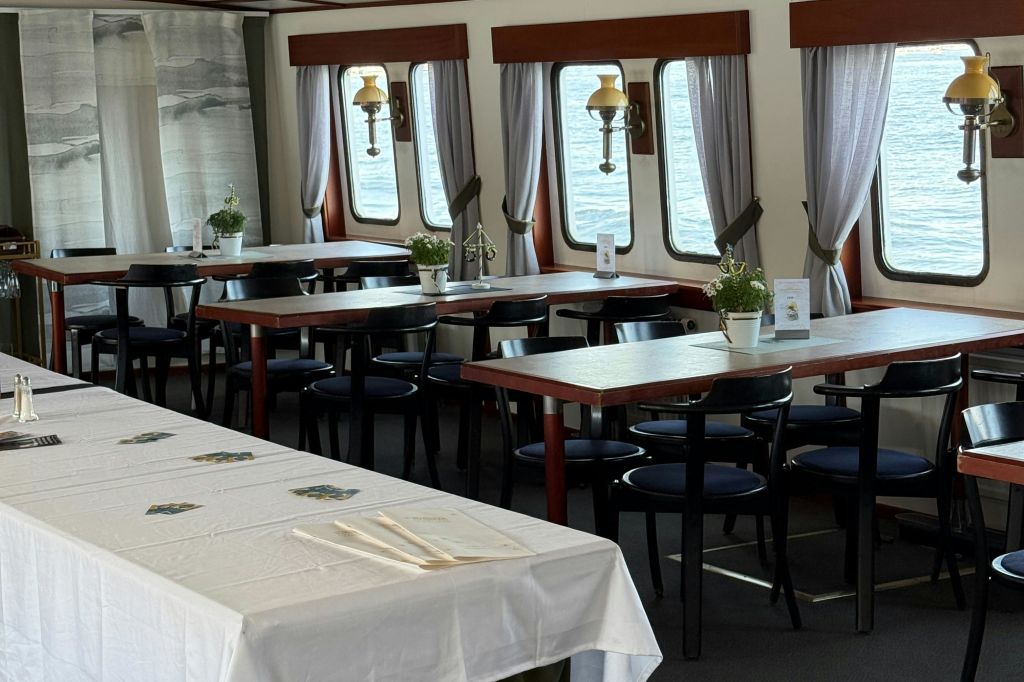 배 위에 여러 개의 테이블과 의자가 놓인 식사 공간으로, 창문에 화분, 메뉴, 커튼으로 장식되어 있습니다.