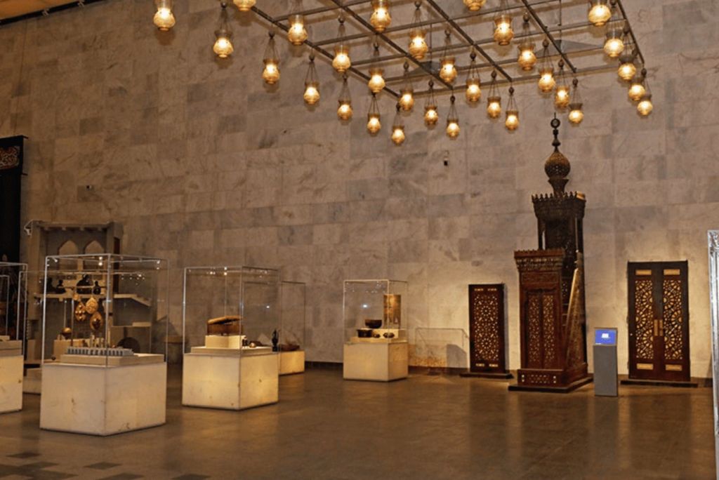 수많은 걸이식 램프가 비추는 유리 케이스와 화려한 나무 조각으로 장식된 유물이 전시된 박물관 전시 공간입니다.