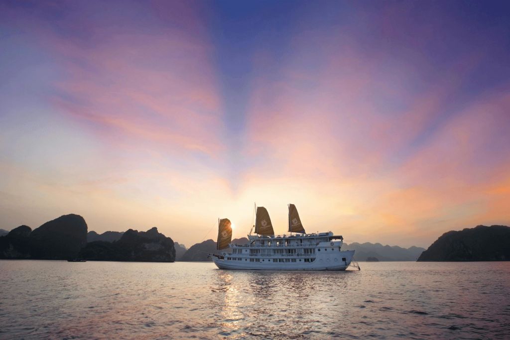 석양이 지면 어두운 돛이 달린 커다란 하얀 배가 잔잔한 바다를 배경으로 산을 배경으로 펼쳐집니다.