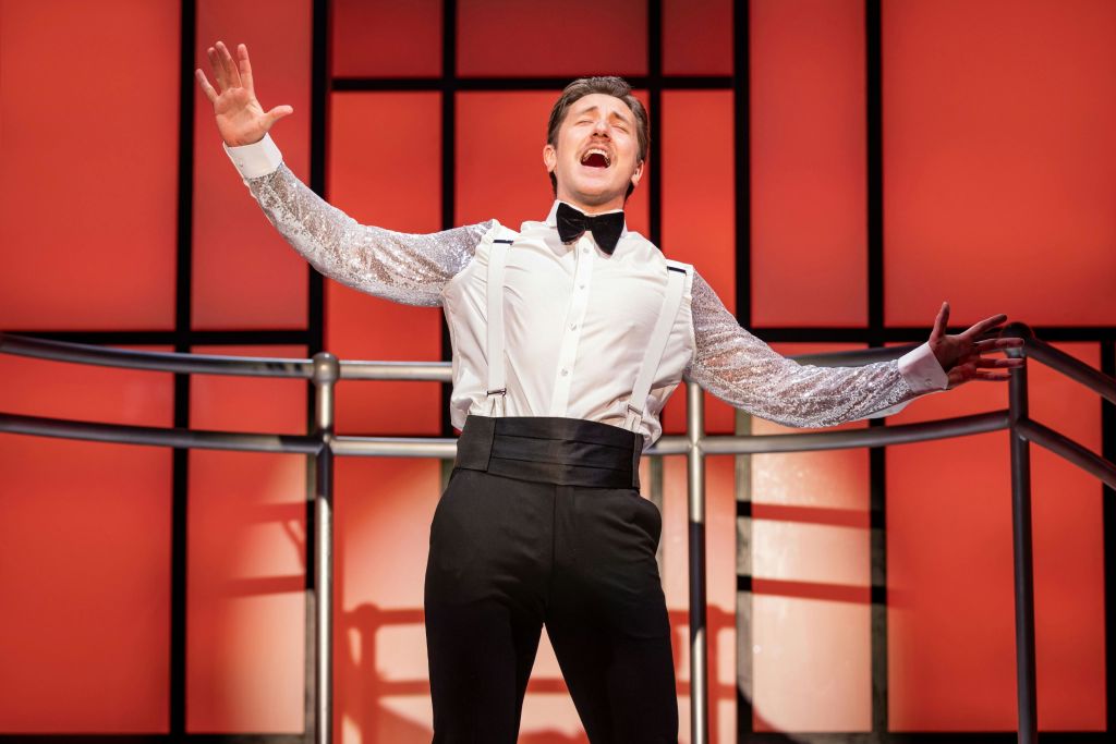 흰색 셔츠와 팔을 쭉 뻗은 나비 넥타이를 입은 남성은 무대에서 빨간색과 검은색 배경으로 열정적으로 노래합니다.