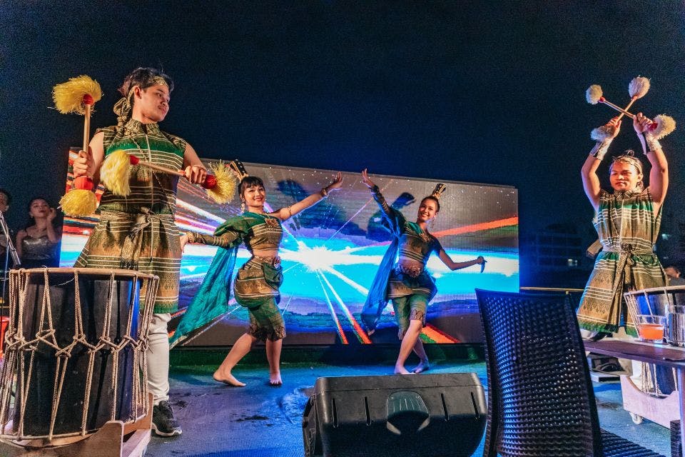 무대에서 전통 복장을 한 세 명의 연주자가 활기찬 디지털 배경으로 커다란 드럼을 연주합니다.