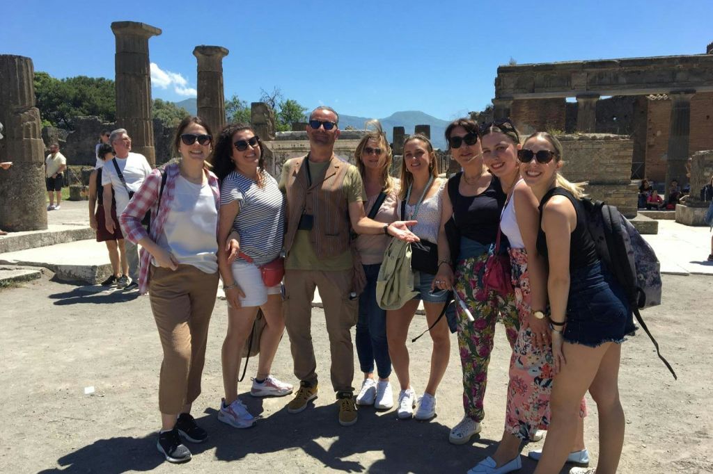 석조 건축물과 맑고 푸른 하늘을 배경으로 햇살이 내리쬐는 고대 유적지에서 포즈를 취하고 있는 사람들.