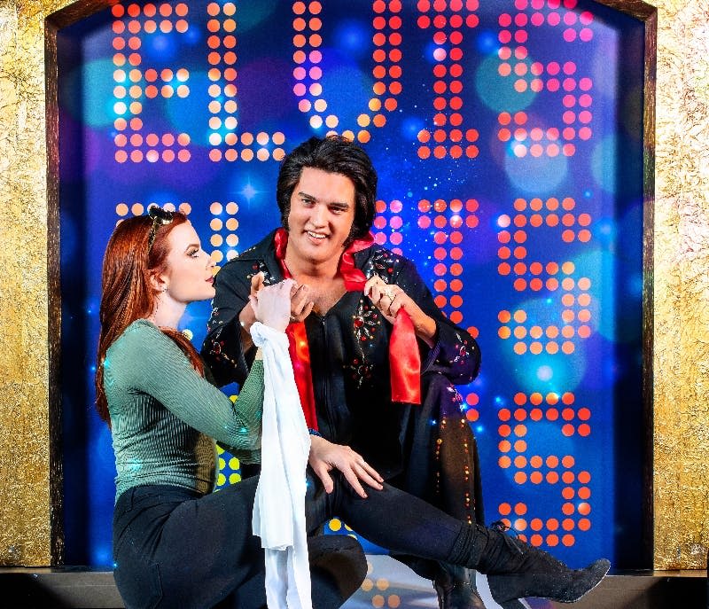 엘비스 복장을 한 남성이 무릎을 꿇고 천을 들고 있으며, 배경에는 화려한 '엘비스' 간판이 있습니다.