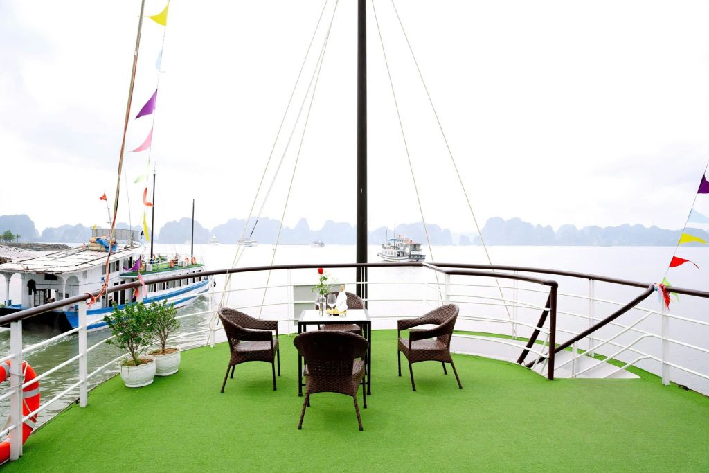 녹색 마루가 깔린 야외 데크, 테이블 주위에 갈색 고리버들 의자 세 개, 화분, 물 위를 배경으로 한 보트.