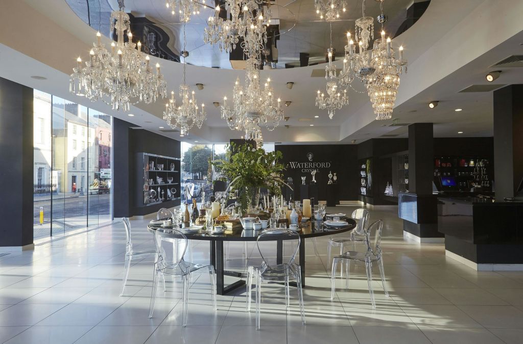 투명한 의자로 둘러싸인 원형 테이블 세트 위에 샹들리에가 걸린 쇼룸입니다. 벽면에 워터포드 로고가 있습니다.