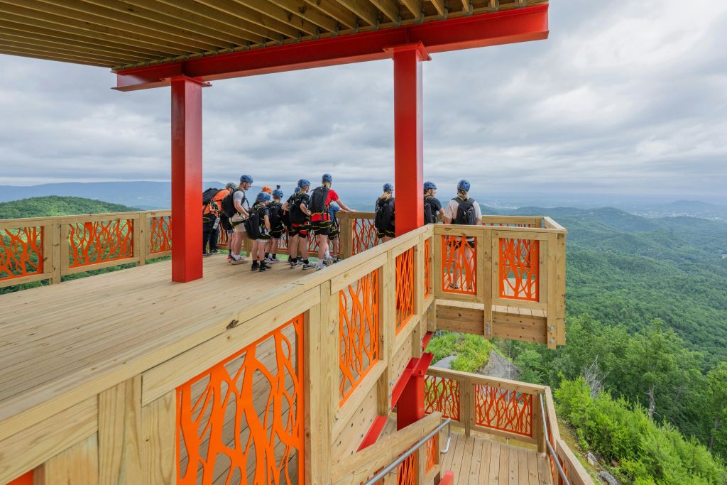 마구와 헬멧을 착용한 한 무리의 사람들이 주황색 난간이 있는 나무 플랫폼에 서서 숲이 우거진 계곡을 내려다보고 있습니다.