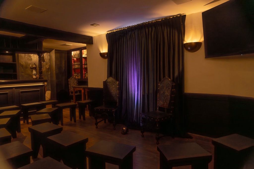 밀폐된 커튼 앞에 화려한 의자 두 개가 있고, 여러 개의 작은 스툴과 테이블이 줄지어 놓여 있는 조명이 희미한 객실입니다.