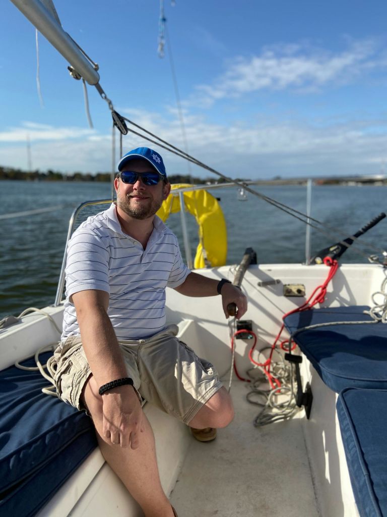 햇살이 내리쬐는 날, 마개와 선글라스를 쓴 남성이 요트에 앉아 물과 육지를 배경으로 사진을 찍고 있습니다.