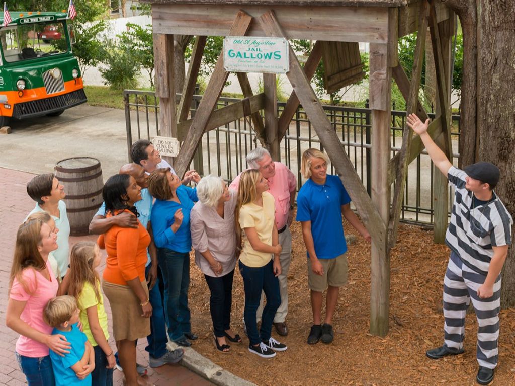 줄무늬 의상을 입은 한 사람이 목조 구조물을 향해 제스처를 취하고 한 무리의 사람들이 세심하게 경청하고 바라봅니다.