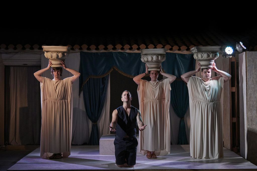 세 명의 배우가 무대에 선다. 한 명은 팔을 펴고 무릎을 꿇고, 다른 한 명은 머리 위에 기둥 모양의 대문자를 들고 서 있다.