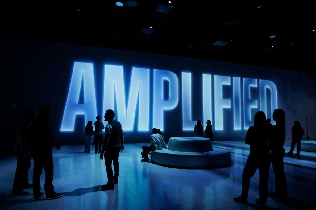 벽에 커다란 조명 "증폭" 표지판이 있는 조명이 희미한 방에 있는 사람들. 어떤 사람들은 서 있고 다른 사람들은 앉아 있습니다.