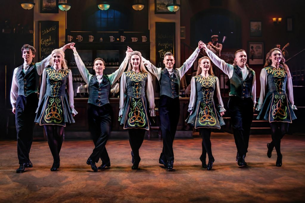 아일랜드 전통 의상을 입은 여섯 명의 무용수가 무대에서 팔짱을 끼고 펍 같은 배경 앞에서 공연을 펼칩니다.