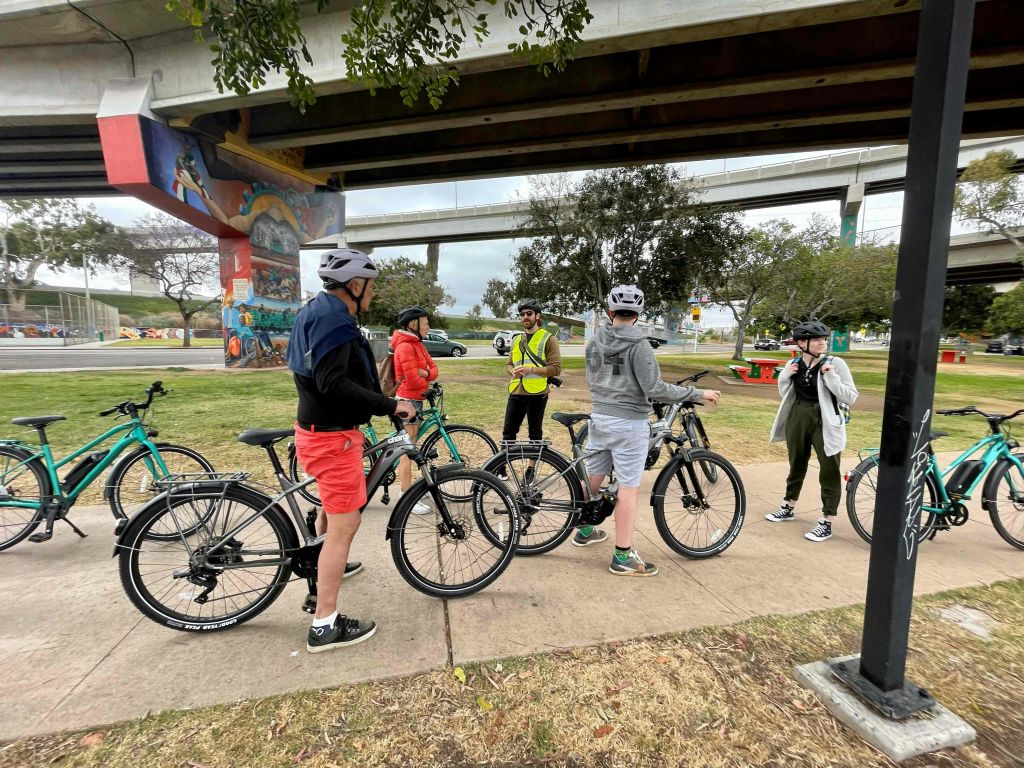 헬멧을 쓴 한 무리의 자전거 운전자들이 다리 아래 모여 자전거와 녹지를 둘러싸고 있습니다. 다리 기둥에 벽화가 보입니다.