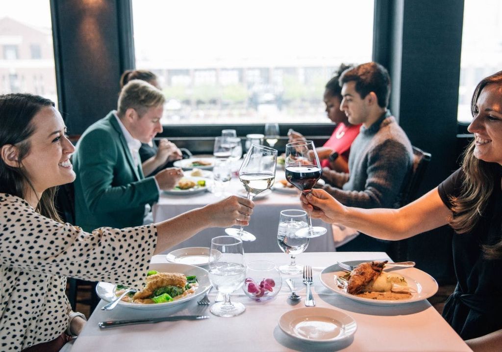 창가 식탁에 앉아 와인잔을 들고 토스트를 하는 사람들, 음식 접시와 식기류를 눈앞에 두고 있는 사람들.