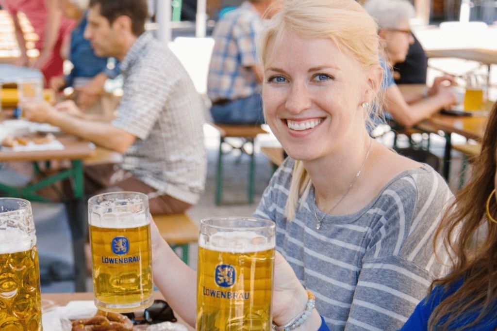 맥주잔 두 개를 들고 있는 한 여성이 야외 테이블에 앉아 미소 짓고 있습니다. 다른 사람들이 배경에 앉아있는 것을 볼 수 있습니다.