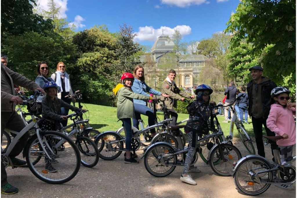 푸른 하늘 아래 푸른 녹음이 우거진 공원의 자전거와 건물을 배경으로 한 어린이를 포함한 일행이 자전거를 타고 있습니다.