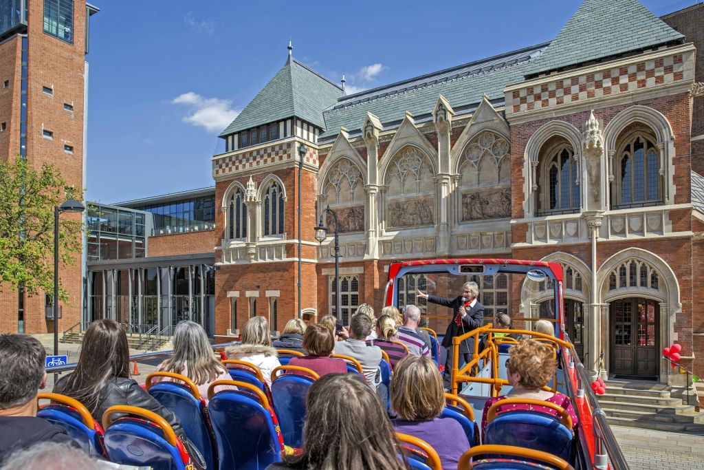 탁 트인 버스에 앉아 맑고 푸른 하늘 아래 화려하고 유서 깊은 건물 앞에서 가이드의 말을 듣고 있는 관광객들.