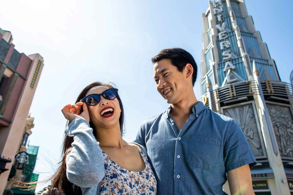 미소를 짓는 두 사람, 선글라스를 쓴 여성과 파란색 셔츠를 입은 남성, "유니버설" 간판이 있는 건물 근처의 야외.