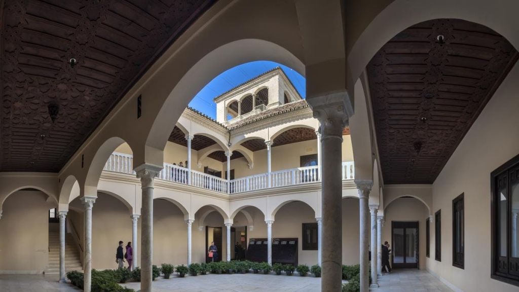 아치형 기둥이 있는 안뜰, 위쪽 발코니, 둘레에 화분이 있고, 아래를 걷고 서 있는 사람들이 있습니다.