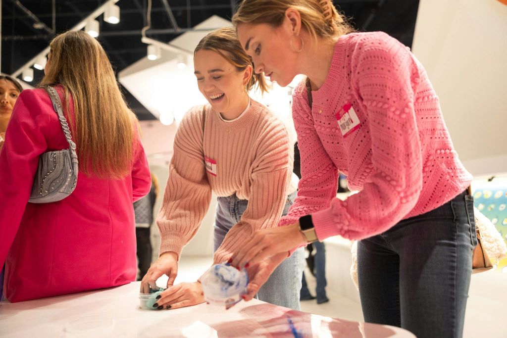 분홍색 스웨터를 입은 두 여성이 미소를 지으며 테이블에서 예술 활동을 하고 있습니다. 배경에는 군중이 보입니다.