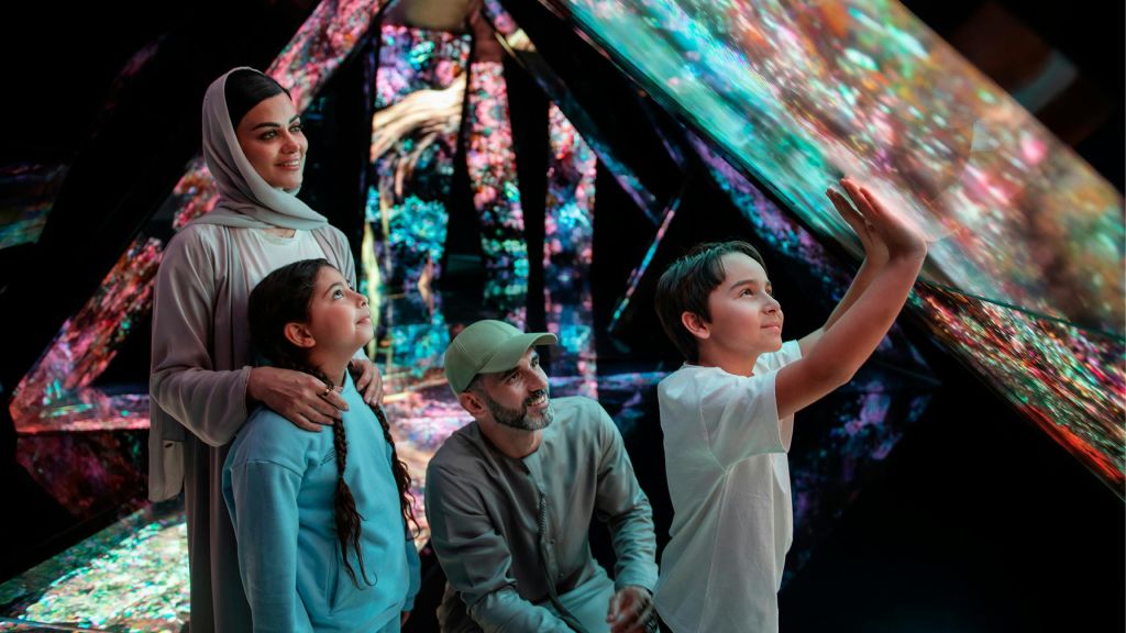 여성, 남성, 두 명의 어린이가 주변 패널에 투사된 디지털 아트와 함께 다채로운 인터랙티브 전시를 탐험합니다.