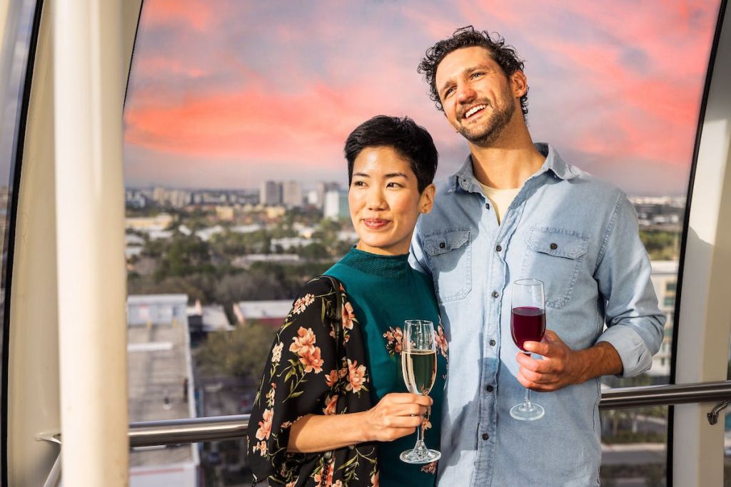 석양이 질 무렵 발코니에 서서 도시의 풍경을 배경으로 한 와인잔을 들고 미소 짓는 커플의 모습입니다.