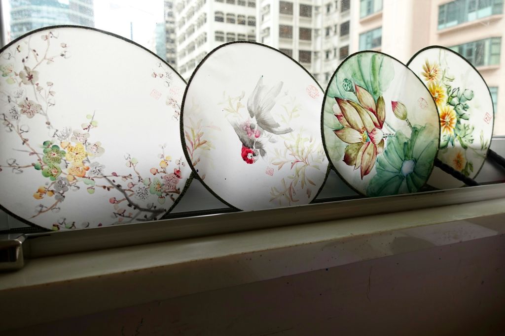꽃과 새 디자인이 돋보이는 5개의 장식용 원형 선풍기가 창턱에 전시되어 있으며, 도시 풍경이 외부에 보입니다.