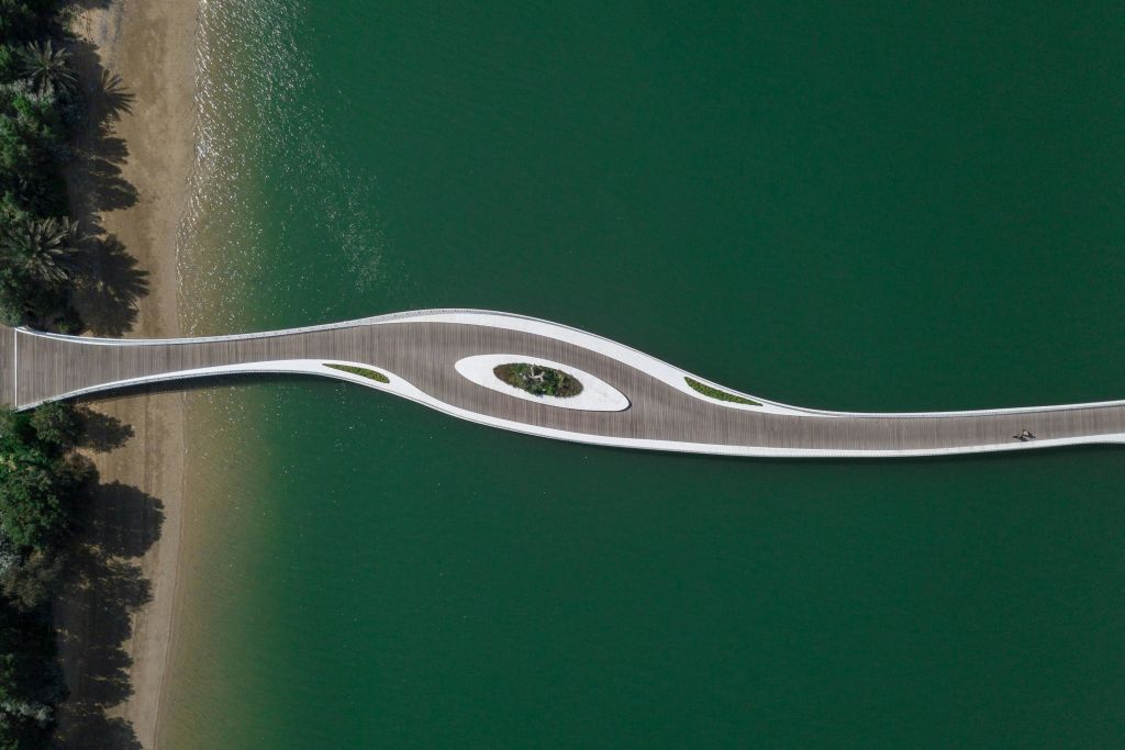 물 위에 녹지가 있는 곡선형의 현대적인 다리의 공중 전망을 감상할 수 있으며, 모래 해변으로 이어집니다.