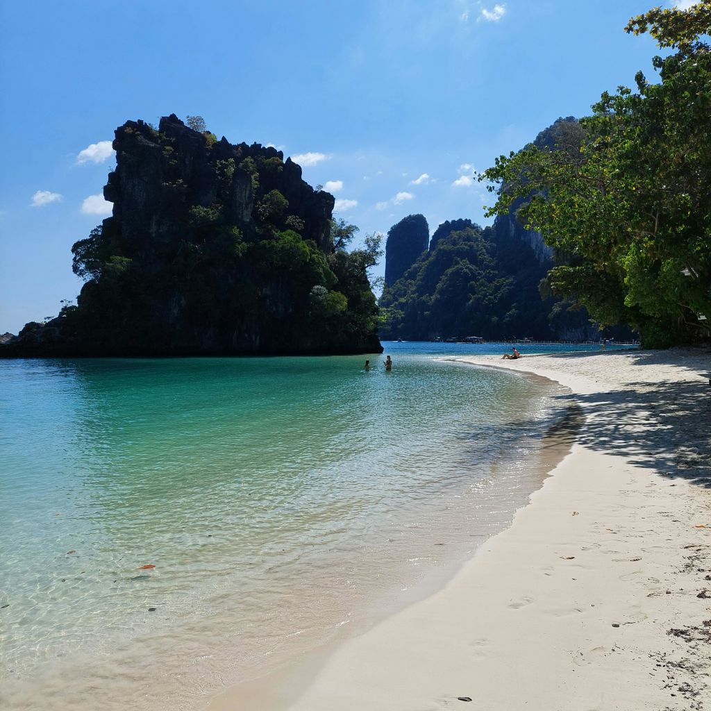 맑은 청록색 바다, 몇 사람이 헤엄치는 모래 해변, 울창한 푸른 나무, 밝고 푸른 하늘 아래 바위 절벽.