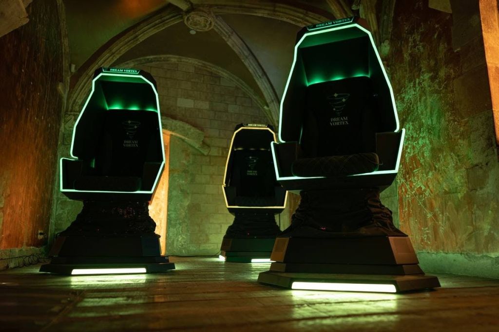 석조 벽과 아치가 있는 희미한 조명의 방에 초록색 가장자리가 빛나는 미래형 검은색 의자 세 개가 있습니다.