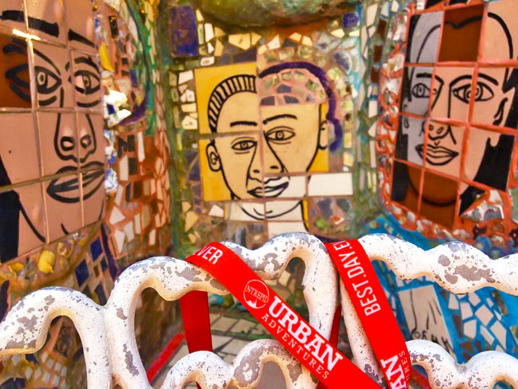 다채로운 초상화와 "어반 어드벤처 (Urban Adventures)" 라고 적힌 빨간색 띠를 들고 있는 흰색 물결 모양 조각의 전경이 있는 모자이크 아트 월.