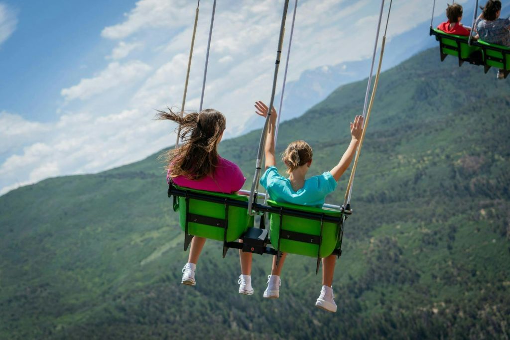 하늘이 맑고 녹음이 우거진 뒤쪽에서 보이는 아름다운 산 풍경 너머로 그네에 앉아 있는 두 사람.