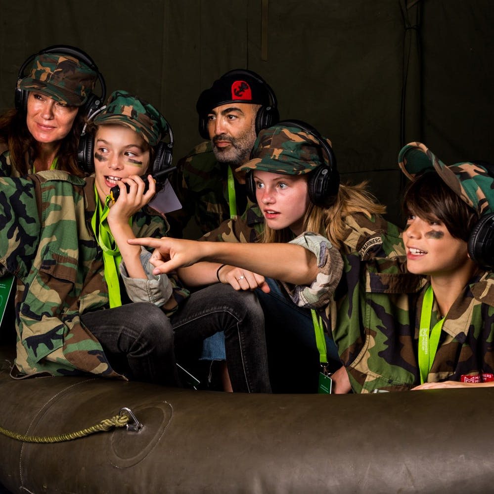 위장복과 헤드셋을 착용한 어린이 4명과 성인 1명이 일렬로 앉아 있습니다. 한 아이는 가리키고 다른 아이는 라디오로 말합니다.