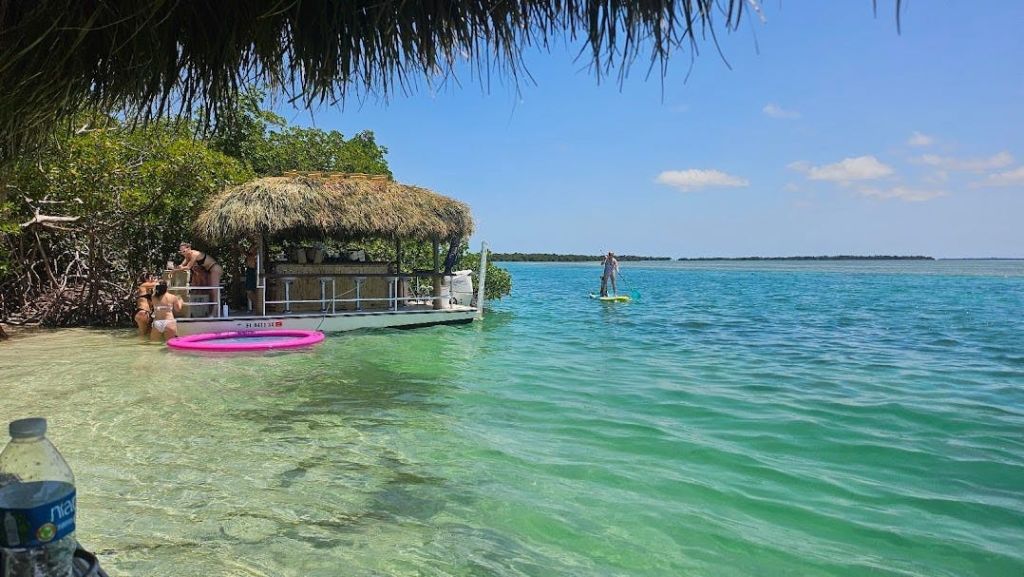 나무를 배경으로 선착장의 초가 지붕 오두막 근처의 맑고 푸른 호수에서 패들보드를 타는 사람.