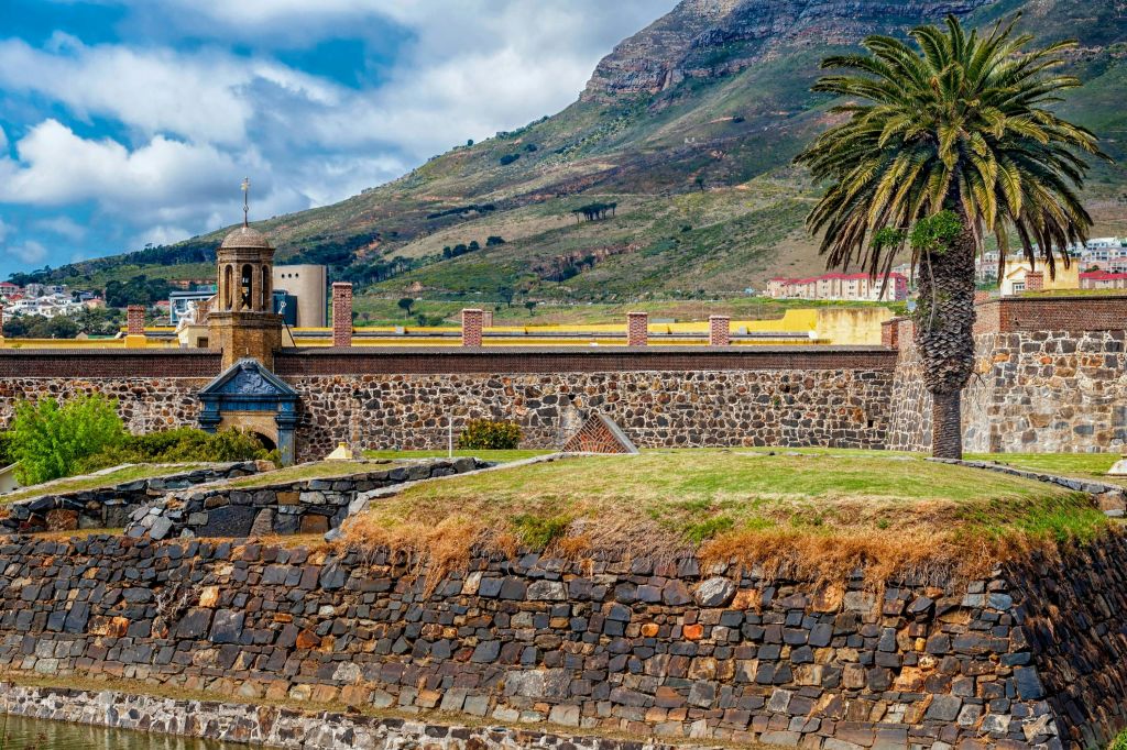 산과 흐린 하늘을 배경으로 잔디가 우거진 꼭대기와 종탑, 야자수가 있는 석조 요새.