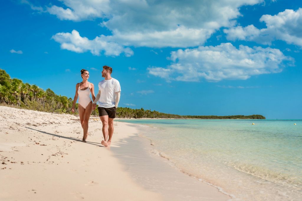 청록색의 맑은 물과 부분적으로 흐린 하늘 아래 야자수가 늘어선 모래사장을 따라 맨발로 걷는 커플.