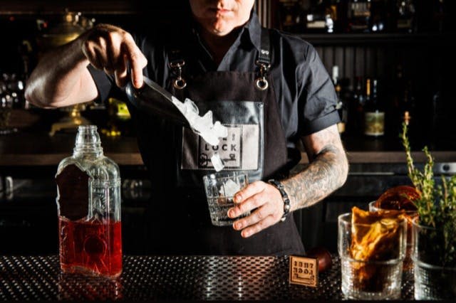 검은색 유니폼을 입은 바텐더가 병과 오렌지를 배경으로 술잔에 얼음을 더하고 있습니다.