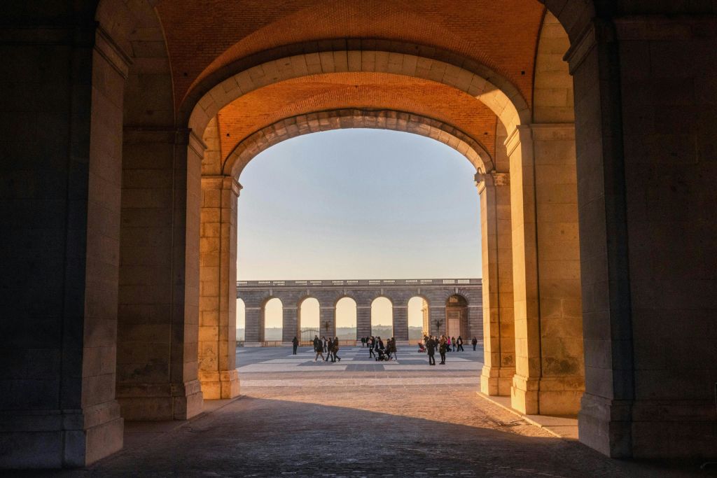 석양을 배경으로 커다란 석조 아치길을 걷고 있는 사람들로, 탁 트인 안뜰과 일련의 아치가 있습니다.