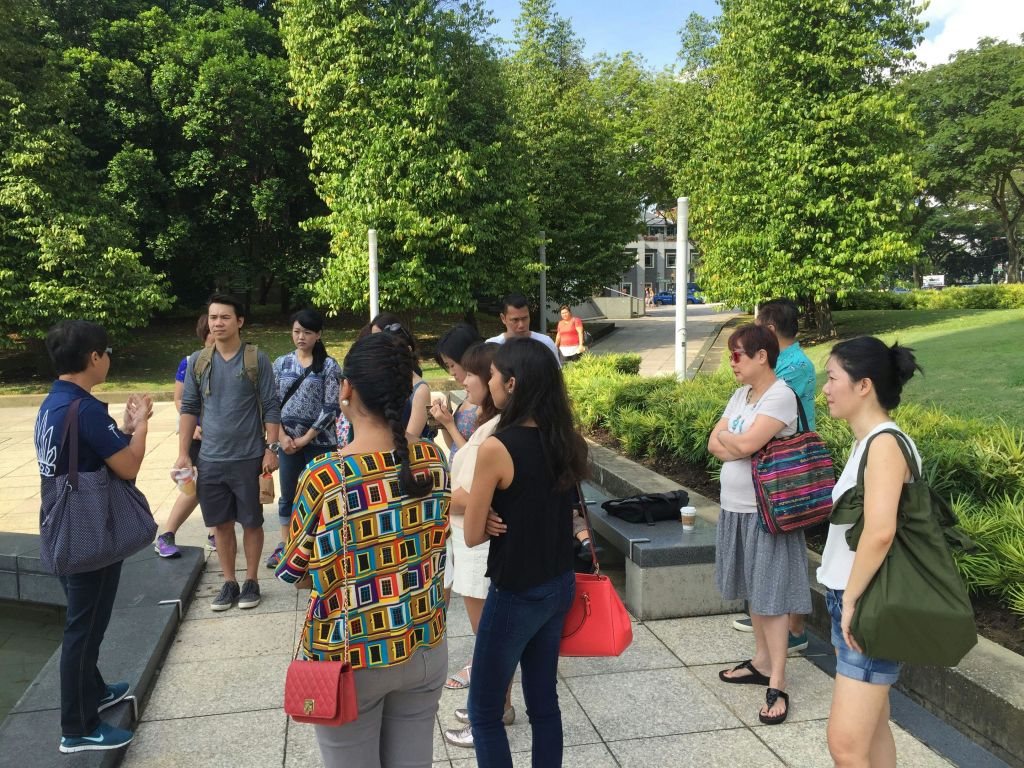 공원에서 야외에 서 있는 사람들, 수다를 떨고 있는 사람들, 주변을 둘러보는 사람들. 녹지와 포장된 길이 보입니다.