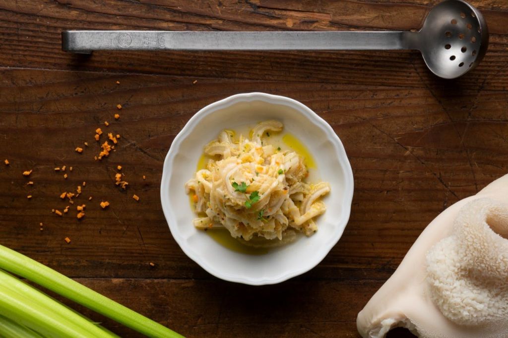 나무 테이블 위에 하얀 그릇에 크리미한 옐로우 소스를 곁들인 파스타가 칼, 셀러리 줄기, 부스러기로 둘러싸여 있습니다.