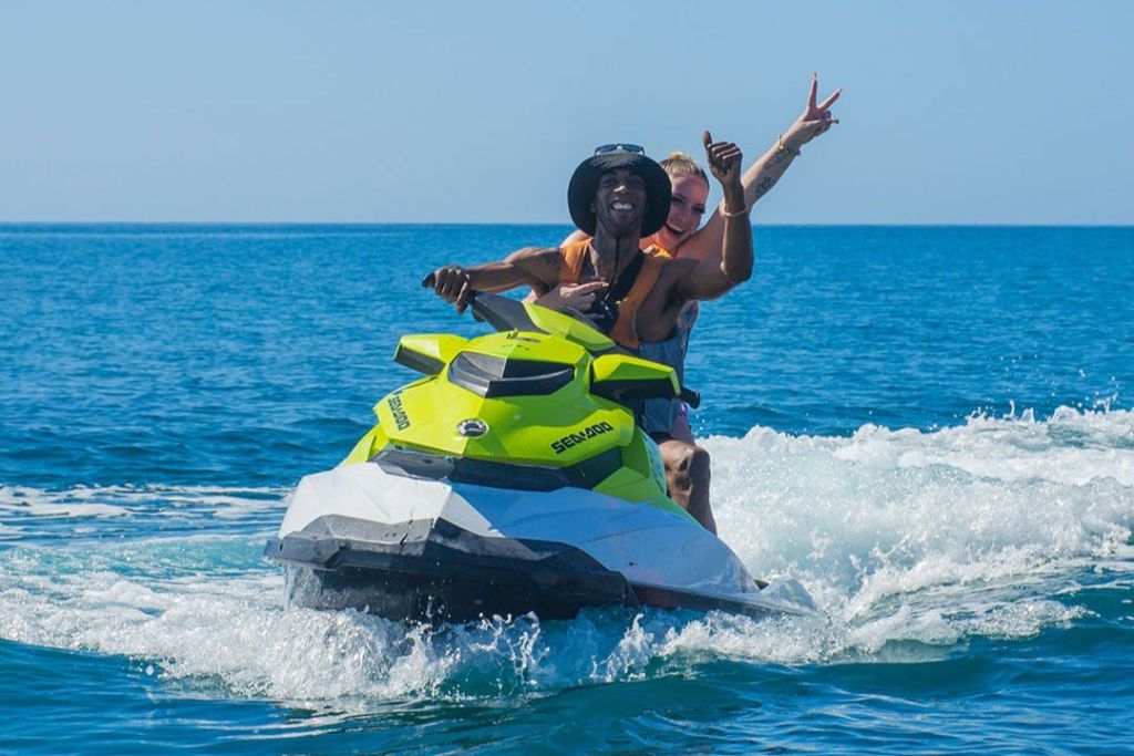 노란색과 흰색 제트 스키를 탄 두 사람이 푸른 물 위에 올라타고 있으며, 승객은 평화 표지판을 만들고 운전자는 미소를 짓고 있습니다.