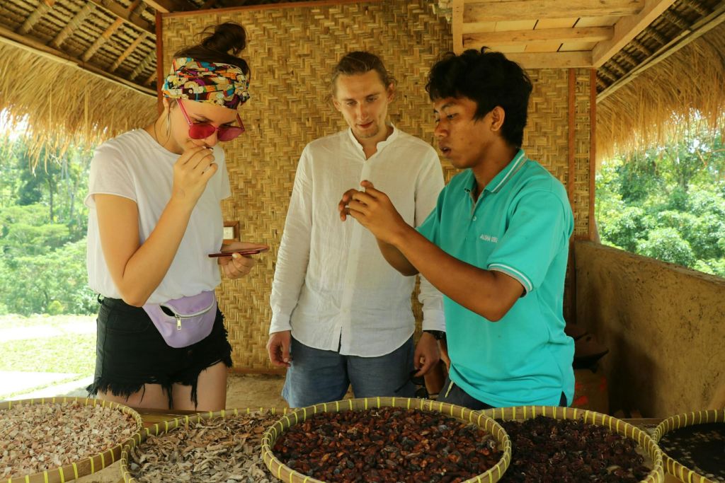 세 사람이 커다란 트레이에 담긴 말린 코코아콩을 소박한 대나무 벽으로 둘러봅니다. 하나는 흰색 셔츠, 다른 하나는 청록색 폴로입니다.